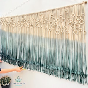 May include: A macrame wall hanging with a gradient of white to green. The top of the hanging features a pattern of circles. The hanging is being trimmed with scissors.