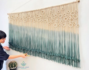 May include: A macrame wall hanging with a gradient of white to green. The top of the hanging features a pattern of circles. The hanging is being trimmed with scissors.