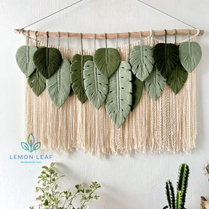 May include: A wall hanging made of macrame with green leaf shapes and a natural wood dowel at the top. The background is a cream colored wall with a white fringe hanging from the bottom of the macrame. The macrame is labeled "LEMON LEAF Handmade by Trash".