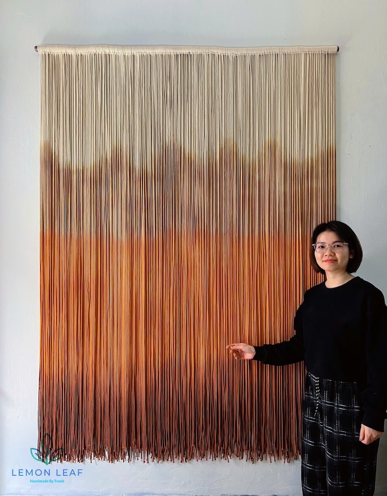 K&ouml;nnte beinhalten: Ein gro&szlig;er Wandbehang aus Makramee mit einem Farbverlauf von Wei&szlig; bis Braun. Der Wandbehang besteht aus vielen F&auml;den, die miteinander verknotet sind. Der Wandbehang h&auml;ngt an einer wei&szlig;en Wand. Eine Person steht vor dem Wandbehang. Der Wandbehang ist mit "LEMON LEAF Handmade by Truth" beschriftet.
