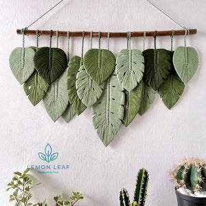 Puede incluir: Un tapiz de pared hecho de hojas de macramé verde, con una barra de madera en la parte superior. Las hojas están dispuestas en forma de cascada, con las hojas más grandes en la parte inferior. El tapiz de pared está etiquetado como "LEMON LEAF Handmade by Trash".