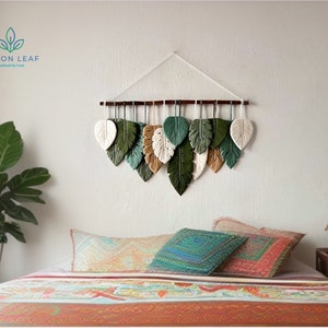 May include: A wall hanging made of woven yarn in the shape of leaves. The leaves are in shades of green, beige, and white. The wall hanging is hung on a white wall above a bed with colorful bedding.