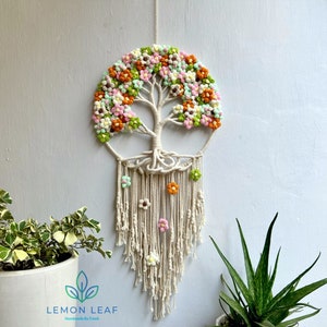 May include: A white macrame wall hanging with a tree of life design. The tree is decorated with colorful yarn flowers in shades of pink, green, orange, and white. The hanging has a fringe of white yarn.