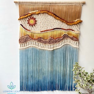 May include: A macrame wall hanging with a sun design in the top section and a blue ombre fringe at the bottom. The sun is made with a yellow, orange, and red yarn. The wall hanging is made with natural colored yarn.