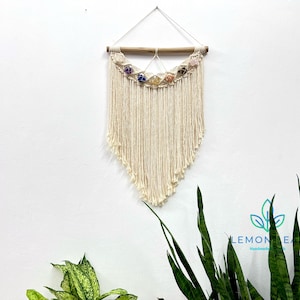 May include: A white macrame wall hanging with a wooden dowel and colorful crystals. The hanging is in the shape of a teardrop and has a fringe at the bottom.