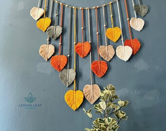 Könnte beinhalten: Ein Wandbehang aus Garn in Form von Blättern. Die Blätter sind in Orange-, Gelb-, Grau- und Weißtönen gehalten. Die Blätter sind mit Perlen aufgereiht und hängen an einer Schnur. Der Wandbehang befindet sich an einer blauen Wand. Der Text "LEMON LEAF" ist in der linken unteren Ecke des Bildes sichtbar.