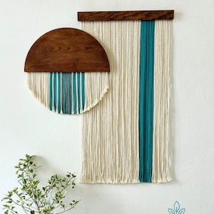 May include: Two wall hangings made of yarn and wood. The larger one is a rectangular shape with a teal stripe down the center. The smaller one is a half-circle shape with teal stripes across the bottom. Both hangings have a natural wood frame.