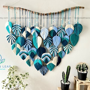 May include: A wall hanging made of macrame with a design of blue and white leaves. The leaves are arranged in a cascading pattern and are attached to a wooden branch. The wall hanging is hung on a white wall with two potted plants in the foreground.