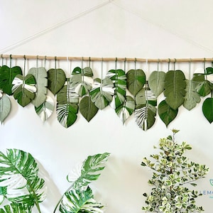 May include: A wall hanging made of green and white yarn, shaped like leaves, with a wooden dowel at the top. The hanging is made of multiple leaves, each with a different pattern. The hanging is called "Lemon Leaf" and is handmade by "Your Name".