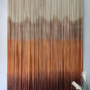 K&ouml;nnte beinhalten: Ein Wandbehang aus Garn mit einem Farbverlauf von Wei&szlig; bis Braun. Das Garn ist zu einer Reihe von Knoten zusammengebunden, um einen strukturierten Look zu erzeugen. Der Wandbehang ist ein beliebtes Dekorationsobjekt f&uuml;r Zuhause.