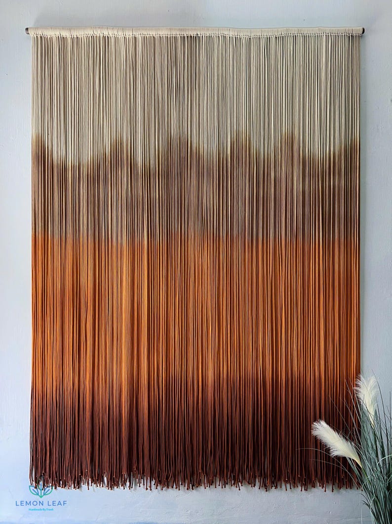 K&ouml;nnte beinhalten: Ein Wandbehang aus Garn mit einem Farbverlauf von Wei&szlig; bis Braun. Das Garn ist zu einer Reihe von Knoten zusammengebunden, um einen strukturierten Look zu erzeugen. Der Wandbehang ist ein beliebtes Dekorationsobjekt f&uuml;r Zuhause.