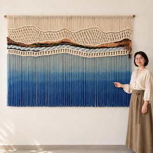 May include: A large macrame wall hanging featuring a landscape design with cream-coloured knotted cord at the top, followed by horizontal bands of brown, green, and grey textures, and a long, dense fringe that fades from light blue to deep navy blue.