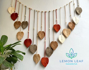 Könnte beinhalten: Ein Wandbehang aus Makramee mit einer Reihe herzförmiger Blätter in Braun-, Beige- und Rottönen. Die Blätter sind an einer weißen Schnur mit Holzperlen aufgefädelt. Der Wandbehang ist mit "LEMON LEAF Handmade By Tranh" beschriftet.
