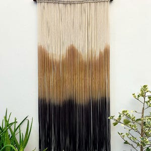 Könnte beinhalten: Ein Makramee-Wandbehang mit einem Farbverlauf aus beige, braunen und schwarzen Garn. Der Behang ist an einer Holzstange befestigt.