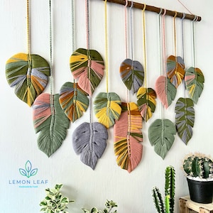May include: A wall hanging made of yarn in the shape of leaves. The leaves are in shades of green, yellow, pink, and gray. The leaves are hanging from a wooden dowel rod. The wall hanging is labeled "LEMON LEAF Handmade by Trudi".