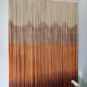 K&ouml;nnte beinhalten: Ein Wandbehang aus Makramee mit einem Farbverlauf von Beige, Braun und Orange. Der Behang besteht aus d&uuml;nnen Schn&uuml;ren und ist an einer Holzleiste befestigt.