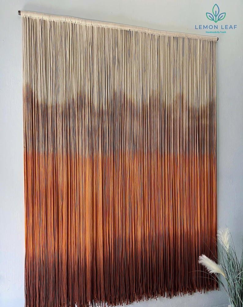 K&ouml;nnte beinhalten: Ein Wandbehang aus Makramee mit einem Farbverlauf von Beige, Braun und Orange. Der Behang besteht aus d&uuml;nnen Schn&uuml;ren und ist an einer Holzleiste befestigt.