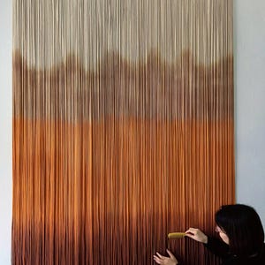 K&ouml;nnte beinhalten: Ein gro&szlig;er Wandbehang aus Makramee mit einem Farbverlauf von Wei&szlig; bis Braun. Der Behang besteht aus vielen einzelnen Garnstr&auml;ngen, die miteinander verknotet sind.