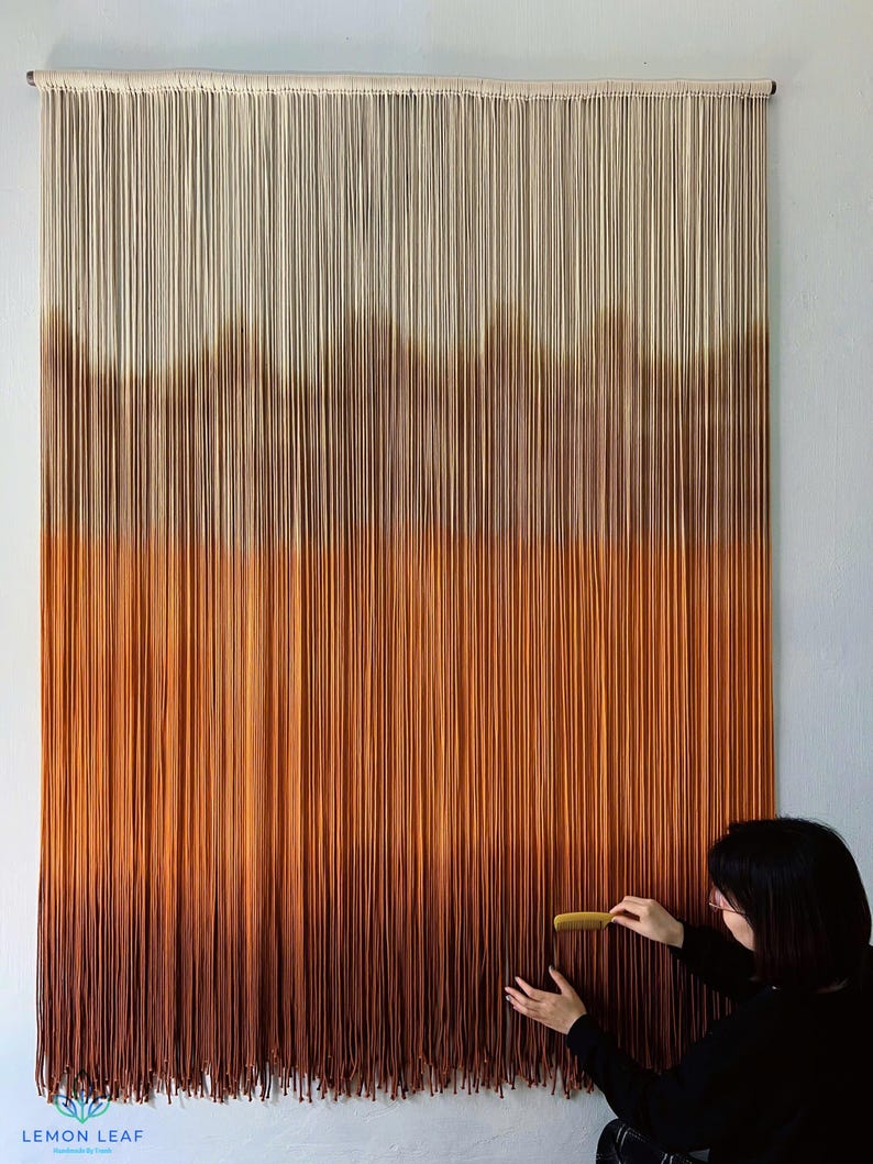 K&ouml;nnte beinhalten: Ein gro&szlig;er Wandbehang aus Makramee mit einem Farbverlauf von Wei&szlig; bis Braun. Der Behang besteht aus vielen einzelnen Garnstr&auml;ngen, die miteinander verknotet sind.