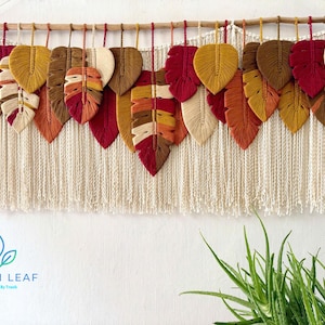 May include: A wall hanging made of woven yarn in the shape of leaves. The leaves are in shades of red, orange, and brown. The hanging has a fringe of white yarn.