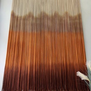 K&ouml;nnte beinhalten: Ein Wandbehang aus Garn mit einem Farbverlauf von Wei&szlig; bis Braun. Das Garn ist zu einer Reihe von Knoten zusammengebunden, um einen strukturierten Look zu erzeugen. Der Wandbehang ist ein beliebtes Dekorationselement f&uuml;r Zuhause.
