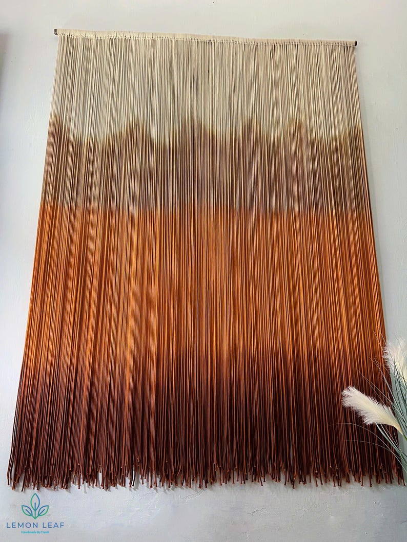 K&ouml;nnte beinhalten: Ein Wandbehang aus Garn mit einem Farbverlauf von Wei&szlig; bis Braun. Das Garn ist zu einer Reihe von Knoten zusammengebunden, um einen strukturierten Look zu erzeugen. Der Wandbehang ist ein beliebtes Dekorationselement f&uuml;r Zuhause.