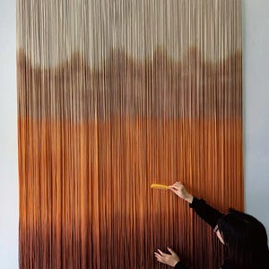K&ouml;nnte beinhalten: Ein gro&szlig;er Wandbehang aus Garn mit einem Farbverlauf von Wei&szlig; bis Braun. Das Garn ist in einer Reihe von Knoten zusammengebunden, um einen strukturierten Effekt zu erzielen. Der Wandbehang h&auml;ngt an einer wei&szlig;en Wand.