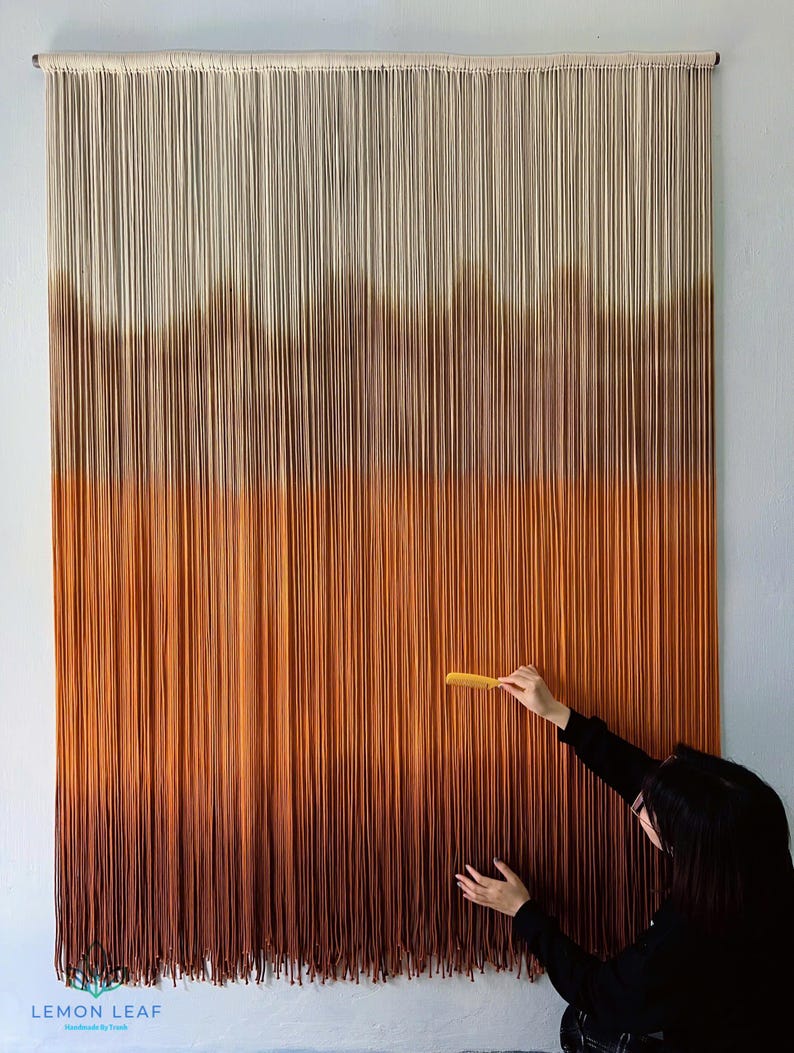 K&ouml;nnte beinhalten: Ein gro&szlig;er Wandbehang aus Garn mit einem Farbverlauf von Wei&szlig; bis Braun. Das Garn ist in einer Reihe von Knoten zusammengebunden, um einen strukturierten Effekt zu erzielen. Der Wandbehang h&auml;ngt an einer wei&szlig;en Wand.