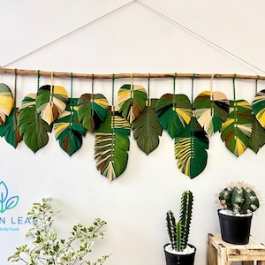 May include: A handcrafted wall hanging featuring 12 large, stylized leaf shapes in shades of green, yellow, and brown. The leaves are suspended from a natural wooden branch with green and yellow cords. The artwork is designed to bring a touch of nature indoors.