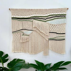 May include: A large, beige macrame wall hanging with a wooden dowel. The design incorporates textured patterns, fringe, and accents of green and black. This handcrafted textile art piece adds a bohemian touch to any space.