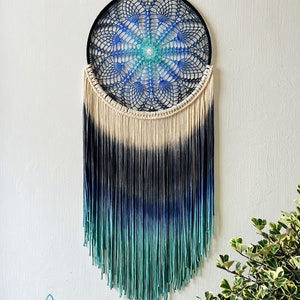 May include: A blue, green, and white ombre dreamcatcher with a crocheted mandala design. The dreamcatcher is hanging from a white wall with a green plant in the corner.