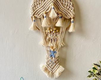 May include: A beige macrame hot air balloon wall hanging with a small doll figure hanging from the bottom. The balloon has a wooden ring and blue and beige tassels.