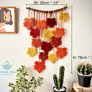 Pode incluir: Uma tapeçaria de macramé de parede com tema de outono. Apresenta uma cascata de folhas de bordo laranja, vermelho e amarelo com um ramo de madeira no topo. A tapeçaria mede 50 cm de largura e 78 cm de altura.