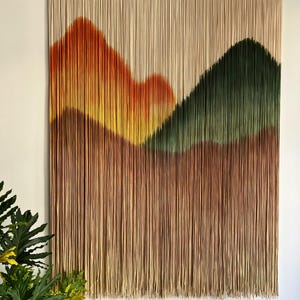 May include: A large wall hanging featuring a mountain landscape design. The artwork uses vertical strands in shades of brown, orange, yellow, and green to create the mountain shapes. The piece is mounted on a wooden bar and is a decorative home accent.