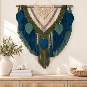 Pode incluir: Uma grande tapeçaria de macramé suspensa numa vara de madeira, com cordões de algodão em camadas nas cores creme, verde sálvia, azul-petróleo escuro e cinzento. O design inclui nós intrincados, secções trançadas, detalhes em folha e borlas.