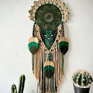 May include: A green and beige macrame dream catcher with feathers hanging from the bottom. The dream catcher is hanging from a white wall with two small potted cacti in the foreground.