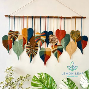 Puede incluir: Un tapiz de pared hecho de hilo de colores, con una serie de hojas en forma de corazón en varios tonos de verde, azul, naranja, rojo y marrón. Las hojas están enhebradas en una varilla de madera y cuelgan de una pared blanca. El texto "LEMON LEAF Handmade by Truth" es visible en la parte inferior de la imagen.
