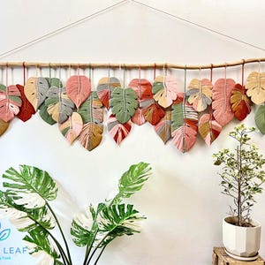 May include: A wall hanging made of woven yarn in the shape of leaves. The leaves are in shades of pink, green, brown, and beige. The leaves are hanging from a wooden branch. The wall hanging is a popular home decor item.