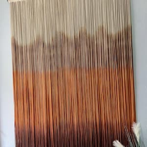 K&ouml;nnte beinhalten: Ein Wandbehang aus Garn mit einem Farbverlauf von Wei&szlig; bis Braun. Das Garn ist oben an einem Holzstab befestigt und h&auml;ngt in langen Str&auml;ngen herunter.