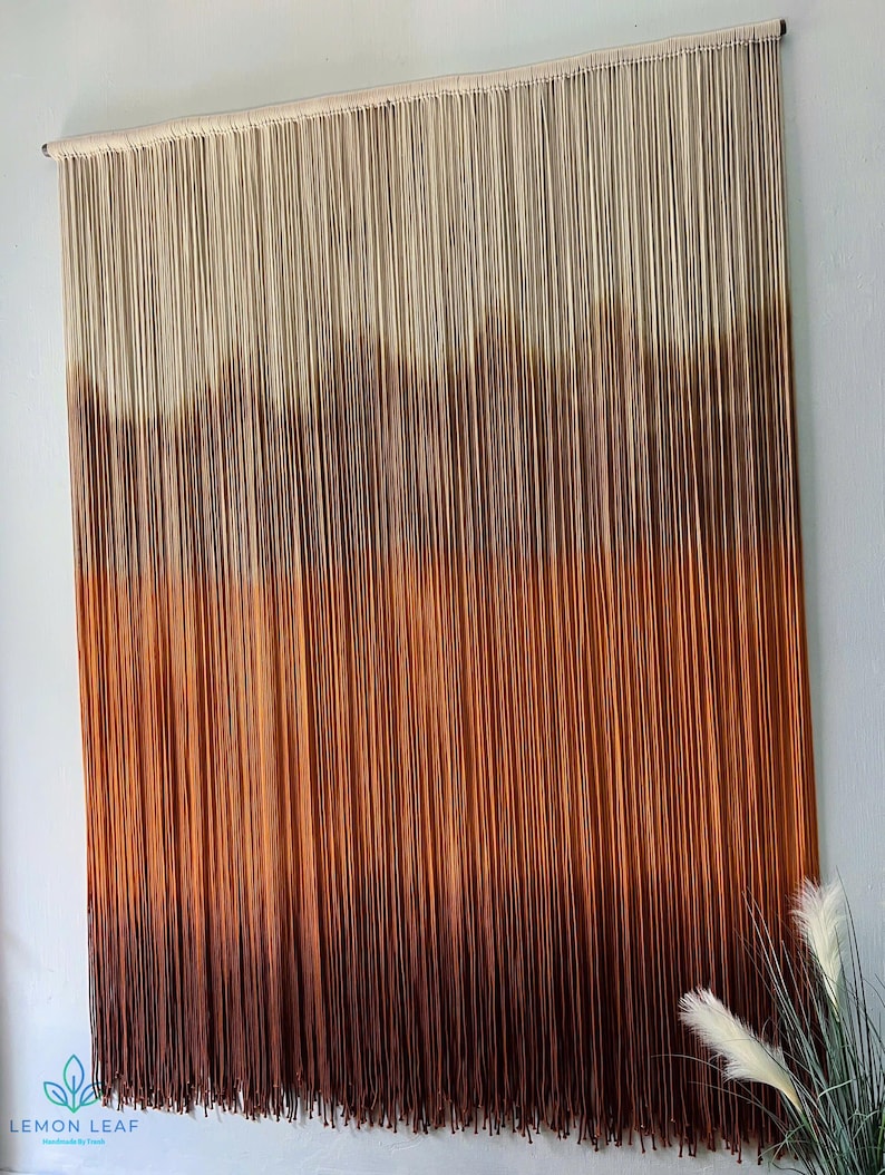 K&ouml;nnte beinhalten: Ein Wandbehang aus Garn mit einem Farbverlauf von Wei&szlig; bis Braun. Das Garn ist oben an einem Holzstab befestigt und h&auml;ngt in langen Str&auml;ngen herunter.