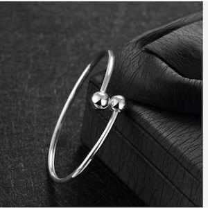 May include: A silver-toned open cuff bracelet featuring a thin, polished metal band that terminates at each end with a smooth, spherical ball bead. The bracelet has a circular shape with a small gap between the two spheres at the opening.
