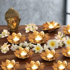 Puede incluir: Una estatua de cabeza de Buda de color dorado colocada junto a varios portavelas de metal dorado con forma de flor de loto, cada uno con una vela de té encendida, intercalados con flores blancas sobre una superficie de madera oscura.