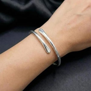 May include: A silver-toned metal cuff bracelet with a bypass design, featuring two smooth, rounded ends that overlap slightly. The bracelet has a polished, reflective surface and a simple, open-ended structure that wraps around the wrist.