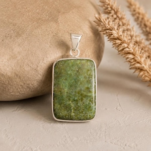 May include: A rectangular pendant featuring a textured, mottled green stone set in a polished silver frame. The pendant includes a silver bail at the top for attachment to a chain, showcasing natural variations in the green mineral surface.