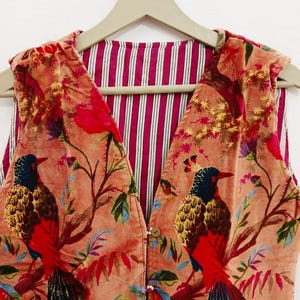 May include: A sleeveless waistcoat featuring a peach-coloured velvet exterior with a repeating pattern of birds perched on branches, accented by red, blue and yellow details. The interior lining displays a vertical striped pattern in pink and white.
