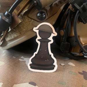May include: A black chess piece sticker with a helmet on top. The sticker is on a camouflage background.