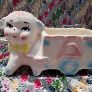 May include: A vintage ceramic planter shaped like a lamb pulling a cart. The lamb is white with pink cheeks, blue ears, and a yellow bow tie. The cart is pink with the letter "A" and blue accents.
