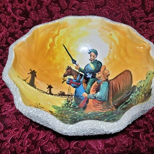 May include: A ceramic bowl with a white rim and a painted scene of two figures on horseback riding through a desert landscape. The bowl has a yellow, orange, and brown background with green and blue accents.