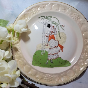 Könnte beinhalten: Ein antiker Keramikteller mit einem erhabenen, geprägten Rand mit Obst- und Traubendesigns. Die Mitte des Tellers zeigt eine farbenfrohe Illustration einer Frau in einem weißen Kleid mit einer roten Schärpe, die in einem grünen Feld mit Bäumen steht.