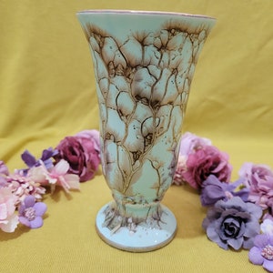 Könnte beinhalten: Eine hellblaue Keramikvase mit braunem Blumendesign. Die Vase hat einen ausgestellten oberen Teil und einen Sockel.